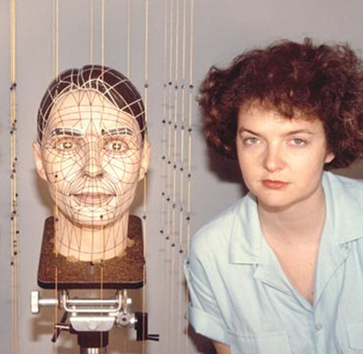 A woman in a blue button up shirt stands next to a model of a human head on a camera stand. Strings with beads hang around the head, which is sectioned off into portions with fractal-like line work.