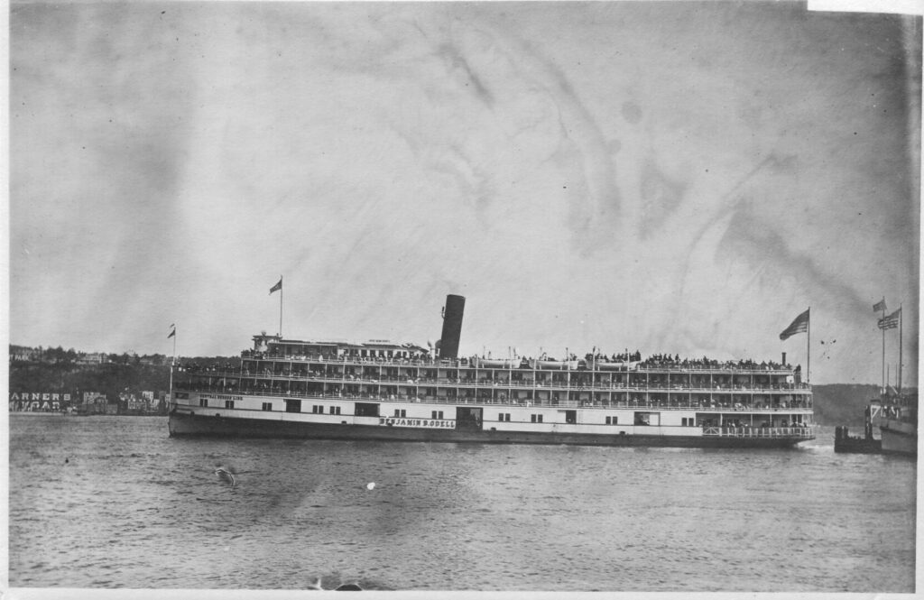 A port-side photograph of the Benjamin B. Odell steamboat as it leaves Manhattan. The ship's top three decks visibly teem with passengers.