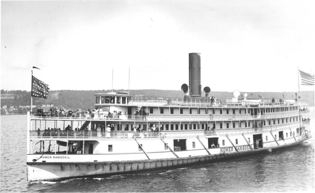 A photograph of the port side of the steamer Homer Ramsdell as it sails along the Hudson River. Dozens of passengers can be seen gathered near the ship's bow, and an American flag is displayed prominently near the stern.