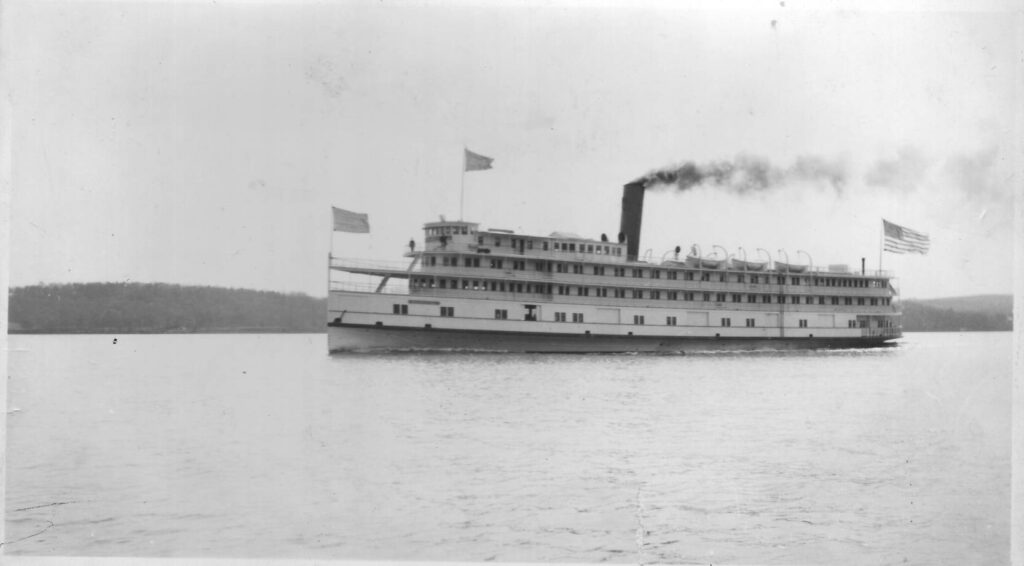 A port-side photograph of the Benjamin B. Odell steamboat on its maiden voyage in April 1911. Black clouds of smoke billow from a smokestack located near in the ship's center. A lonely figure can be seen standing on the top deck near the bow.