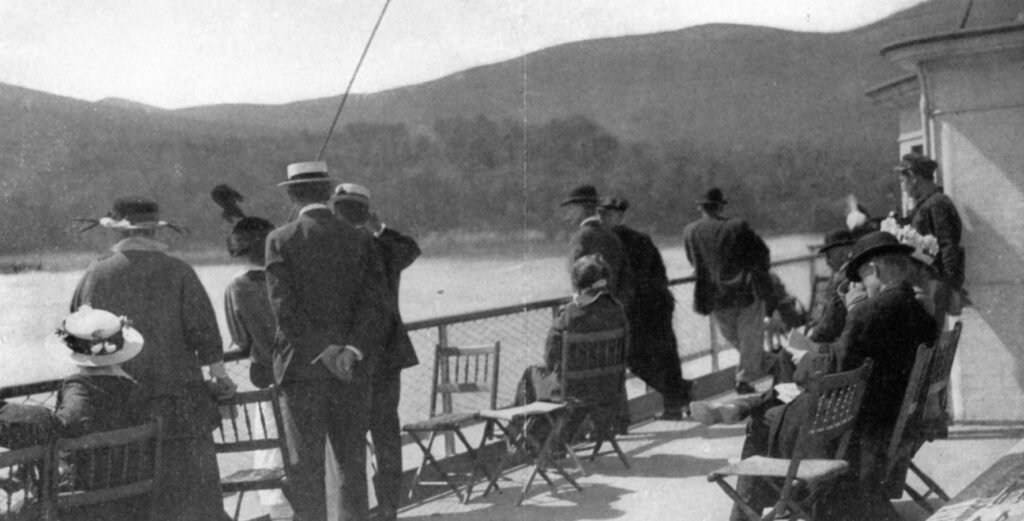 A photograph depicting a group of more than a dozen well-dressed people relaxing, socializing, and taking in the view aboard the deck of the Benjamin B. Odell while it is underway. Some passengers are seated on wooden folding chairs while others are standing. A seated man in the foreground is reading a book.