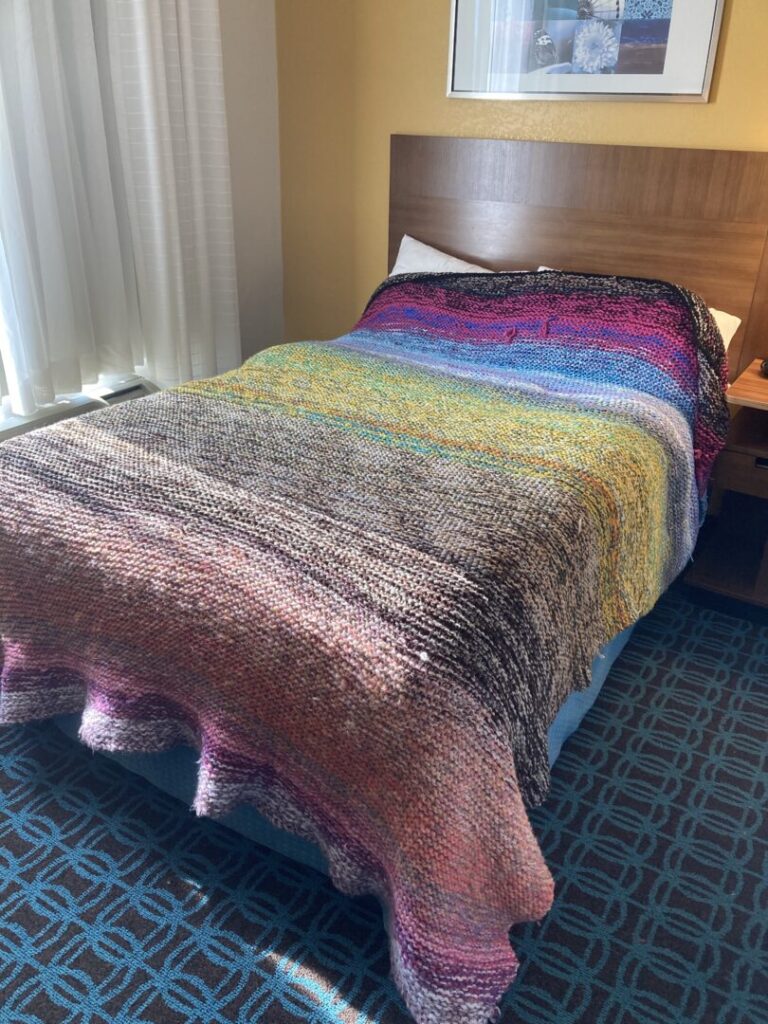 A large, knitted blanket covers a bed in a hotel room with gold walls, teal patterned carpet, and white curtains. The yarn colors shift throughout the blanket, creating a rainbow gradient. Streaks of sunlight beam across the bed.