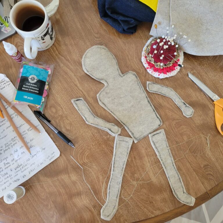 A light tan felt doll they have created. The doll’s body and limbs are disconnected and laid out on Sav’s kitchen table with thread, pens, an open notebook, buttons, a coffee mug, extra fabric, a red velvet pin cushion and sewing scissors are surrounding the doll.
