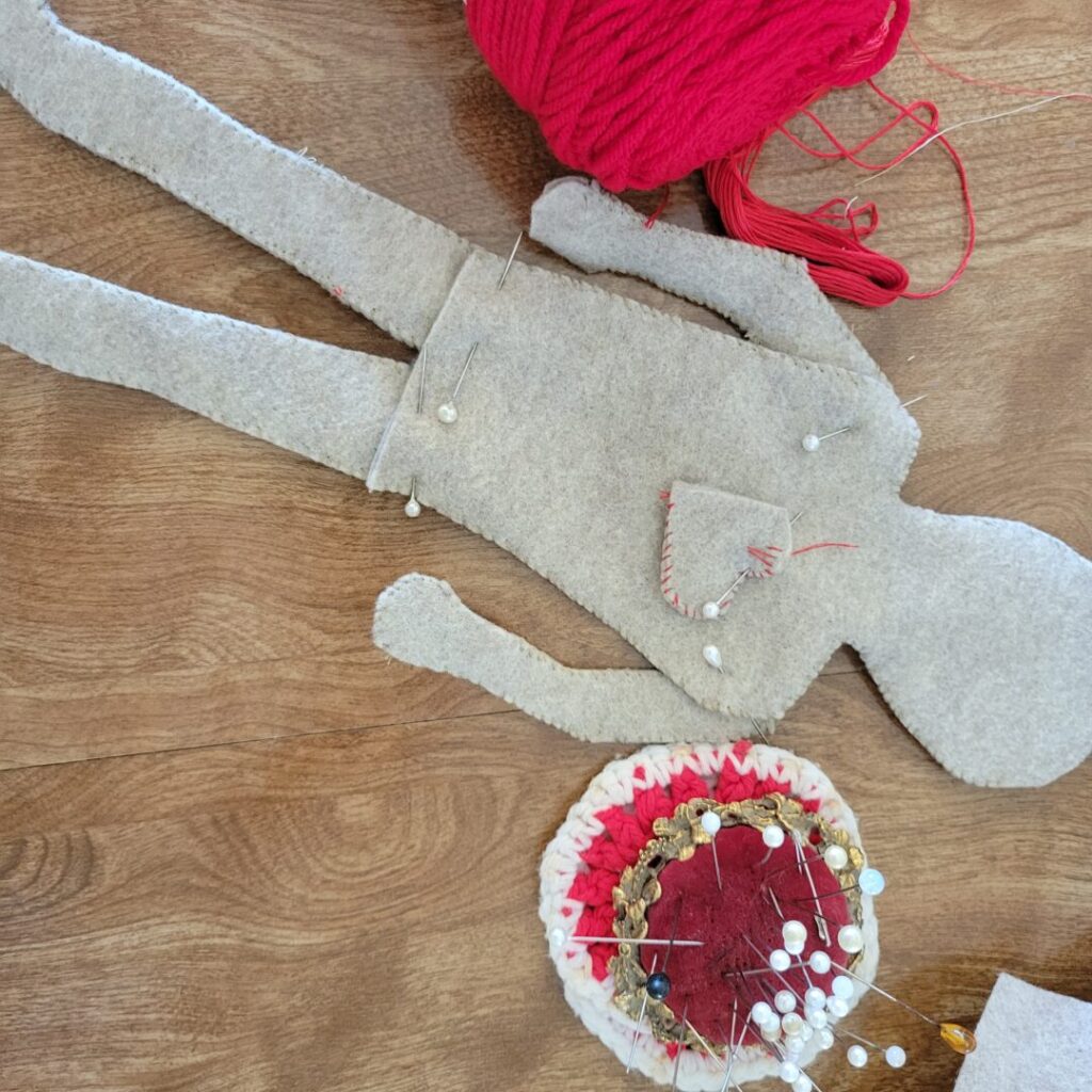 A photo of a light tan felt doll they have created. The limbs and body are pinned together. A red velvet pin cushion and red thread are beside the doll laid out on Sav's kitchen table.