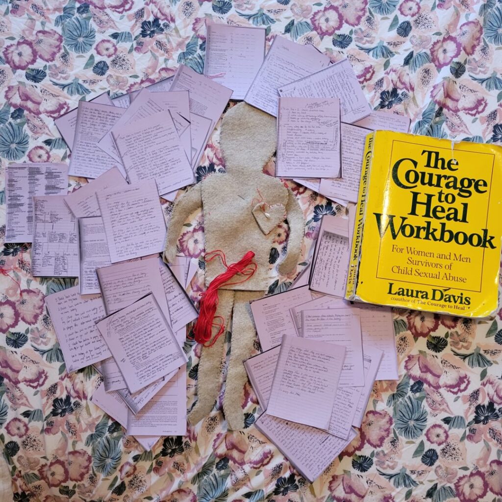 The felt doll is on Sav's bed surrounded by small purple sheets of paper of photocopied pages of "The Courage to Heal." The large bright yellow workbook is next to the doll, and there is red string on top of the doll.