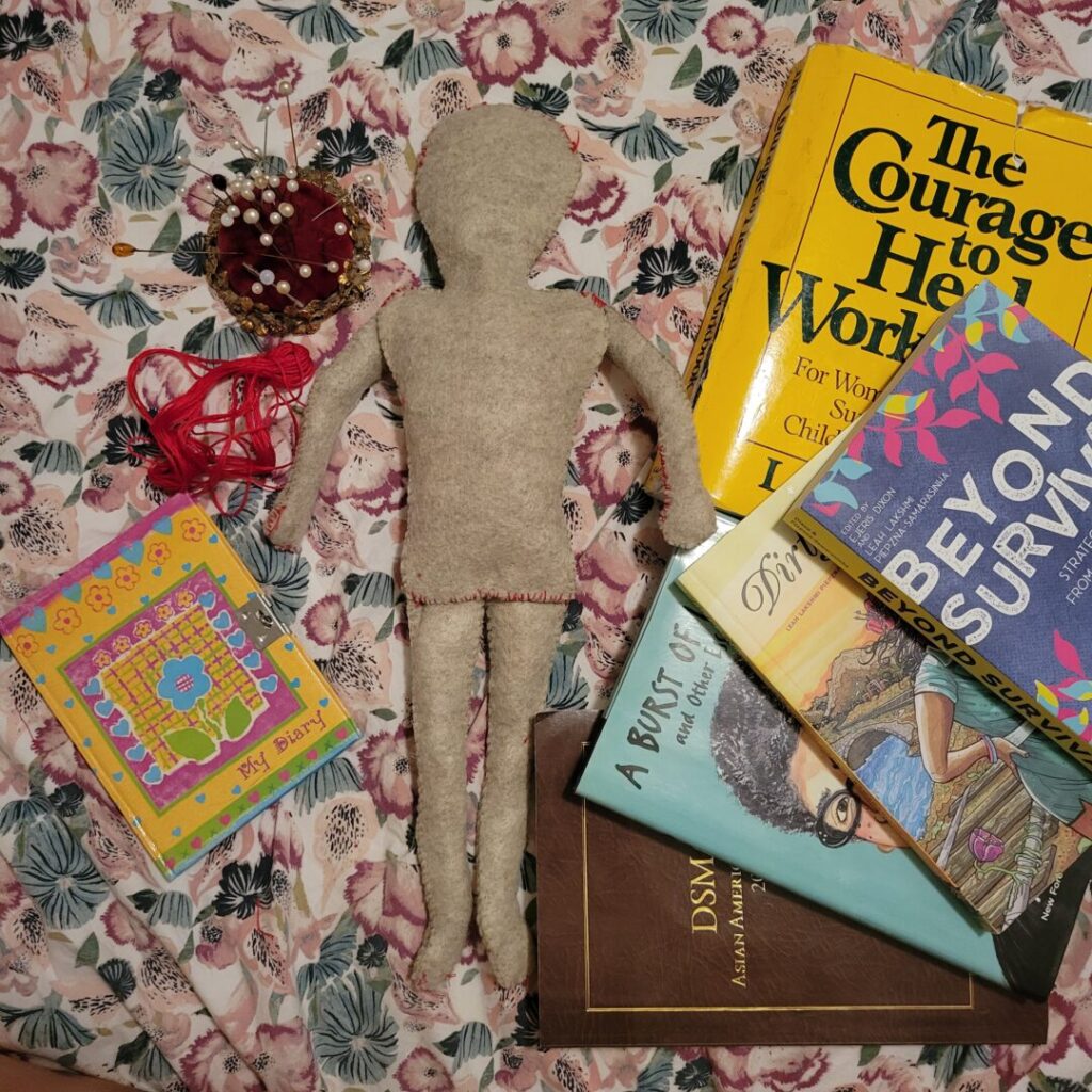 A photo taken by Sav of a light tan felt doll on Sav’s bed, with pink and green floral sheets. Surrounding the doll are Sav’s childhood diary, red thread, a red velvet pin cushion, The Courage to Heal Workbook and several books: Beyond Survival Strategies and Stories from the Transformative Justice Movement, Dirty River, A Burst of Light, and the “hacked” DSM from Open in Emergency