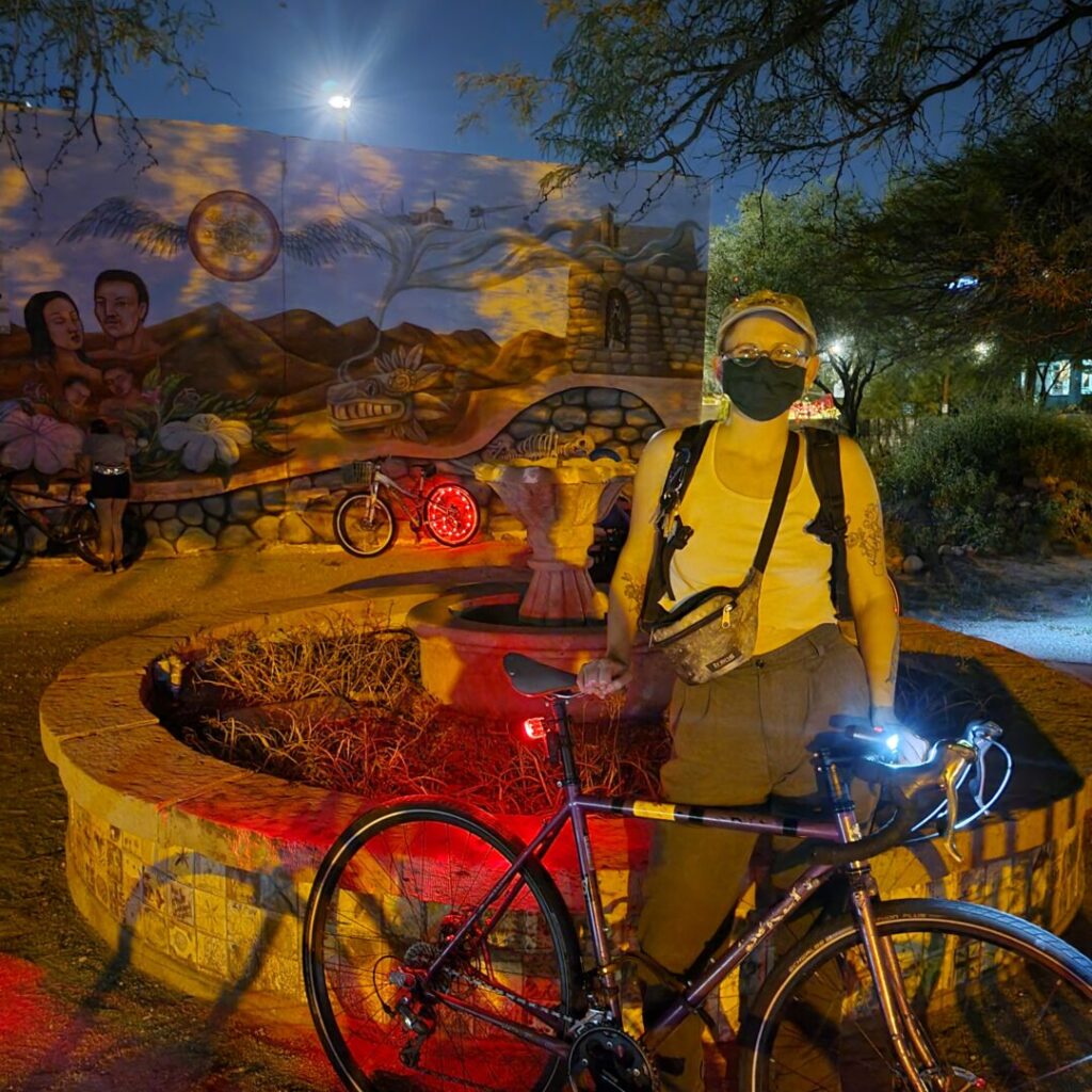 A photo taken of Sav from 2021 from a community ride in Tuscon, Arizona. Sav is standing with their pink sparkly bike. Behind them, there is an old town fountain and a community mural depicting the Sonoran Desert. Sav is wearing a white tank top, brown dress pants, green cloth mask, fanny pack, and baseball hat.