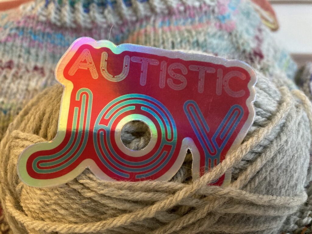 A holographic pink sticker, reading “autistic joy” is stuck in a ball of gray yarn. This sticker is designed by Jen White Johnson. A blurred piece of knitted fabric is in the background of the image.