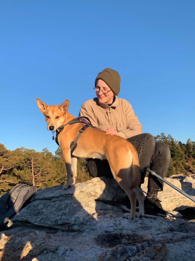 A photo of Sav taken March 2020 on top of Mount Lemmon in Tucson with their dog Cassie. Cassie is a medium tan mixed-breed dog, they are standing on a boulder next to Sav, who is sitting next to Cassie. They are wearing black jeans, a tan jacket, and a green beanie. Their silver cane is positioned between their legs.