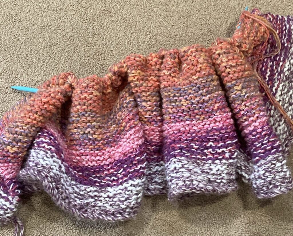 A piece of knitted fabric, about a foot tall, on a blue knitting needle, laying on beige carpet. The colors shift through white, purple, pink, and peach. The knitting is ruffled as it is scrunched up to fit on the length of the needle.