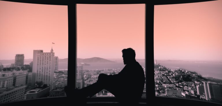 Silhouette of a young man looking out a window of a high-rise apartment onto a cityscape below.