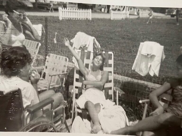 A black and white image of a group of girls in the Catskills; they wear cropped sleeveless tops and shorts. Joyce--possibly 15 years old--holds court in the center, laying on a lounge chair with an arm extended. The girls listen intently, laughing. One stands, one sits in a chair, and one sits in a wheelchair.