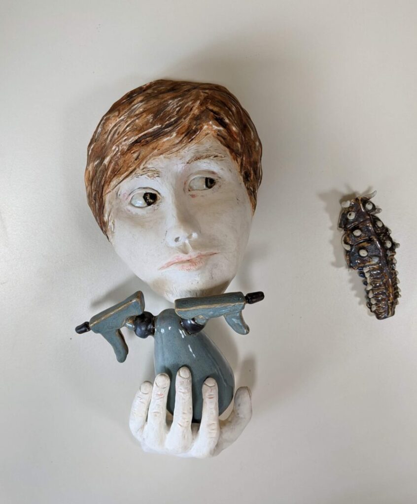 Photo of an art piece made out of clay (ceramic with glaze and acrylic paint). It depicts a man’s head peering to the side at a bug crawling around nearby. The man’s hand cradles a double-headed spray bottle. One side of the spray bottle points to the insect, while the other points off into the distance.