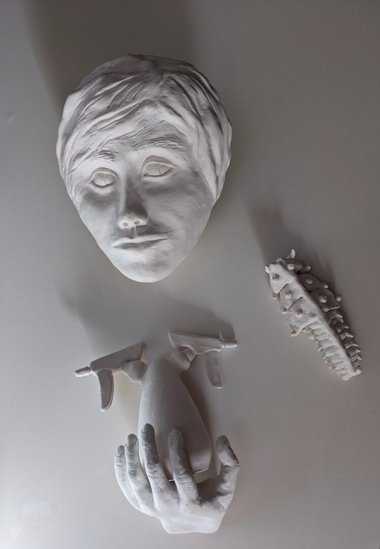 Photo of an art piece made out of clay (ceramic unglazed). It depicts a man’s head peering to the side at a bug crawling around nearby. The man’s hand cradles a double-headed spray bottle. One side of the spray bottle points to the insect, while the other points off into the distance.