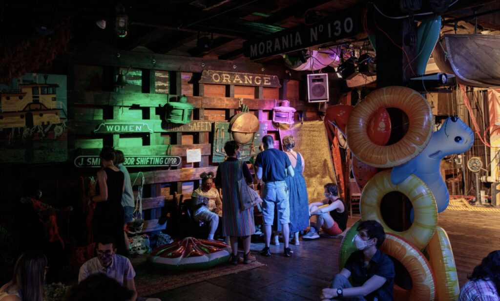 Barge interior quarter view with a mixture of people standing and sitting on chairs, pillows and rugs throughout. The light in the image moves from left to right with low light in the stim fringe joy area to brighter light on the stage area. Wooden walls are covered with wooden signs, lanterns, ropes, bells, and barge paintings. A small wooden boat hangs from the ceiling. A broad wooden pole is covered with inflated pool floats. The floats are brightly colored and range in sizes and shapes, from fruit slices to imaginary and real sea beings. A large brightly colored watermelon slice with a metallic sheen is on the floor for sitting.
