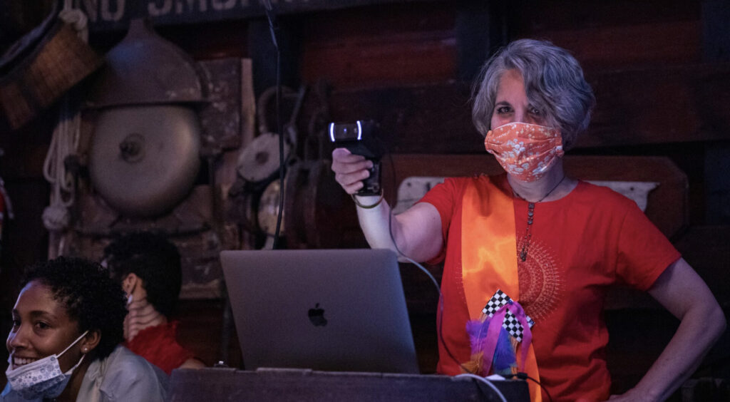 A white woman with short wavy silver black hair and dark eyes looks into the camera with an amused expression. An orange and white printed mask covers her nose and mouth. She stands behind a laptop with her right arm and hand stretched forward holding a camera. Her left arm is crooked at the elbow, her hand resting on her hip. She wears a bright red short sleeve t-shirt with a large, delicate, cream colored screen print. A long multi-strand, intricately beaded necklace dangles over the screenprint. A silky bright orange wide sash drapes from her right shoulder towards her hip. The sash has a black and white checkered rectangle covered in hot pink ribbon and brightly colored feathers on it. To her right is a smiling Black person with short cropped, curly black hair. They have dark brown eyes and wear a white and blue printed mask on their chin and a light blue jacket. Behind them is a white person with short hair and black tattoos on their neck, wearing a red high-collared sleeveless shirt. The background is a rough wooden wall covered in fishing baskets, ship ropes, knots, and pulleys.