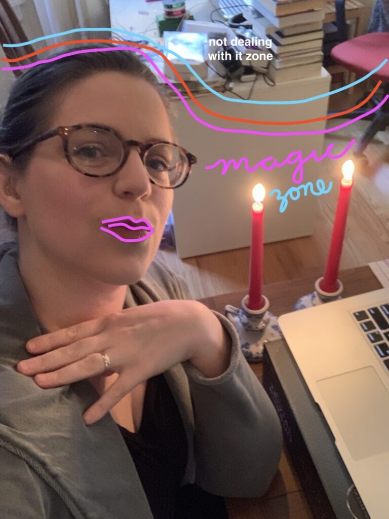 Margaret, a white person with tortoise shell glasses, brown hair, and a gold-glitter earmold wearing a blue-gray hoodie. She is seated at a table with a laptop on top of a board game box, with two red candles lit beside her. In the background is a desk with a pile of books and messy electronics cords. Wavy pink red and aqua lines have been drawn on top of the photo, dividing the desk in the background from the party set-up. The messy desk is labeled "not dealing with it zone" and the rest has been labeled "magic zone" in cursive. Margaret's lips are also outlined in hot pink.