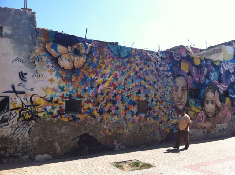 Mural of a small, orange butterfly in the top left, and on the right a picture of a young girl in pink dress, from the waist up, and the face of a middle-aged woman (maybe her mother) in purple headscarf. In between them and the orange butterfly are countless, multi-colored Xs, giving the visual effect of a flock of butterflies at a distance.
