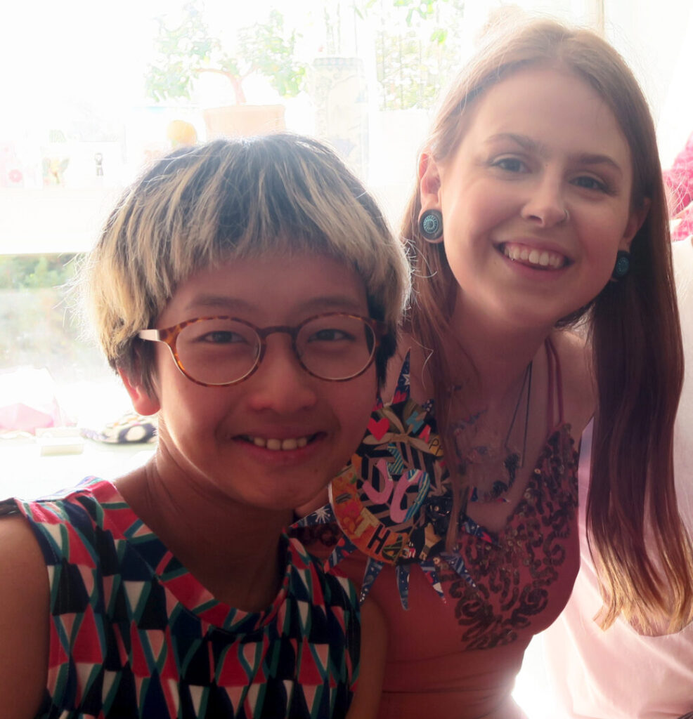 Denise (left) sits next to Sophie (right) at Sophie’s birthday party. Denise is an Asian woman with glasses and highlighted short hair. She is wearing a red, blue, black, and white sleeveless top with a geometric pattern. Sophie is a white woman with long walnut-coloured hair.  She is wearing large blue earrings, a nose ring, a pink sleeveless dress with sequins, and a badge that says “happy 30th birthday”. They are both facing the camera and smiling.