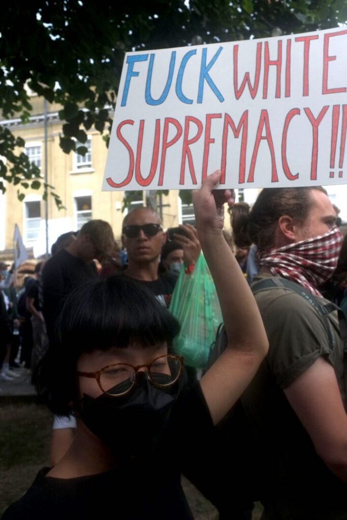 Denise is holding up a placard in her left hand above her head, which says “FUCK WHITE SUPREMACY!!” the lettering for “fuck” is in blue and the lettering for “white supremacy” is in red. Denise is an Asian woman with short black hair and glasses. She is wearing a black short sleeves t-shirt and a black mask that covers most of her face. Denise is stood towards the front of a large crowd of people under a tree with a yellow building in the background.