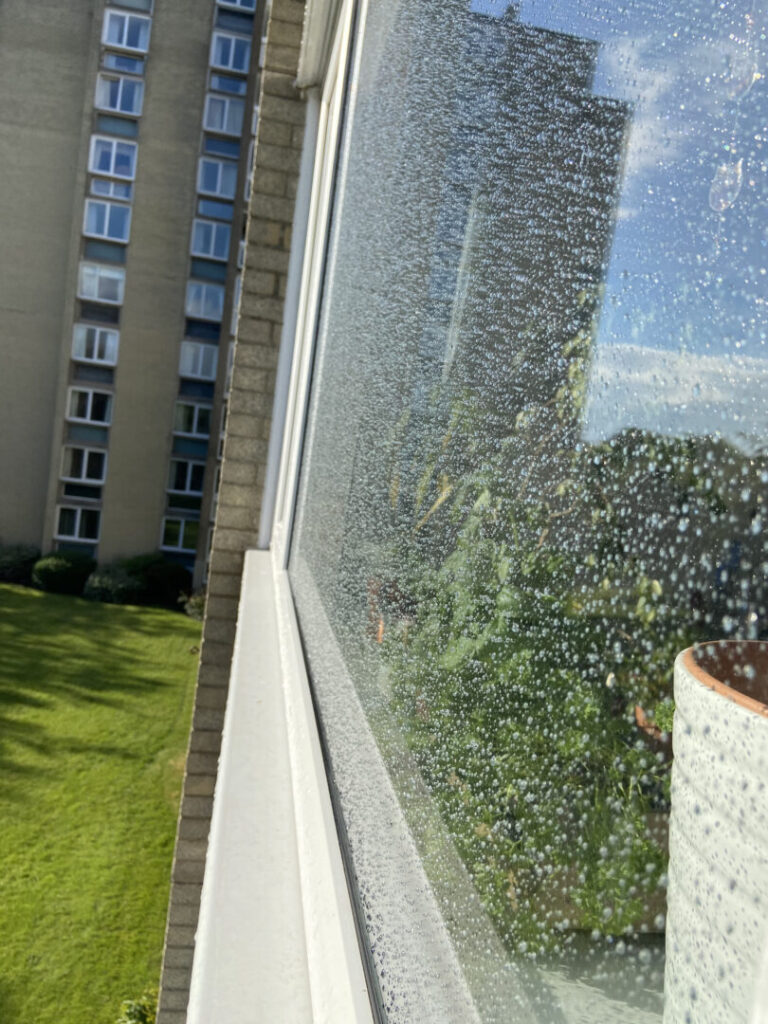 Image of the outside of a window looking in. This horizontal photo was taken leaning out of a window at a ninety-degree angle. The left side of the photograph shows a block of flats and green grass. The right side of the photo consists of the outside of the window which has water droplets on it and it shows the reflection of the building, sky and greenery. On the far right of the photo, there is a plant plot and plants, which are inside.