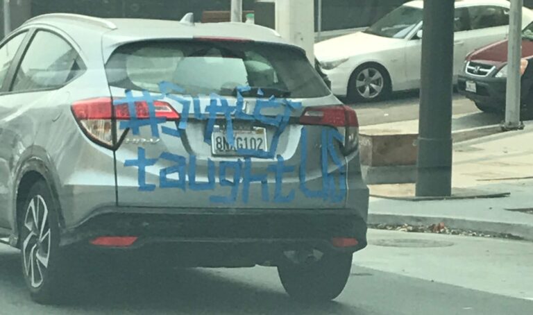 The image of the back of a gray car was taken in San Francisco, California. On the hatchback of the car it says “# Stacey Taught Us” in blue masking tape