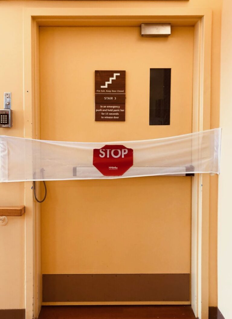 A yellow exit door in a locked dementia unit. A keypad to exit without setting off an alarm is to the left of the door. Across the door is white mesh banner with large red stop sign in the middle. STOP is printed in white capital letters.