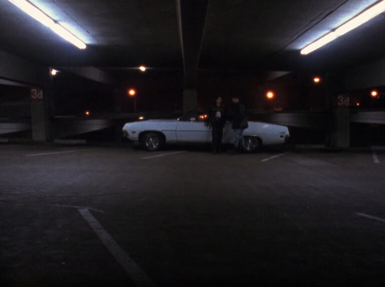 Two people in dark clothing stand against a white convertible in a dark, empty parking garage. They are posed a significant distance away from the camera.