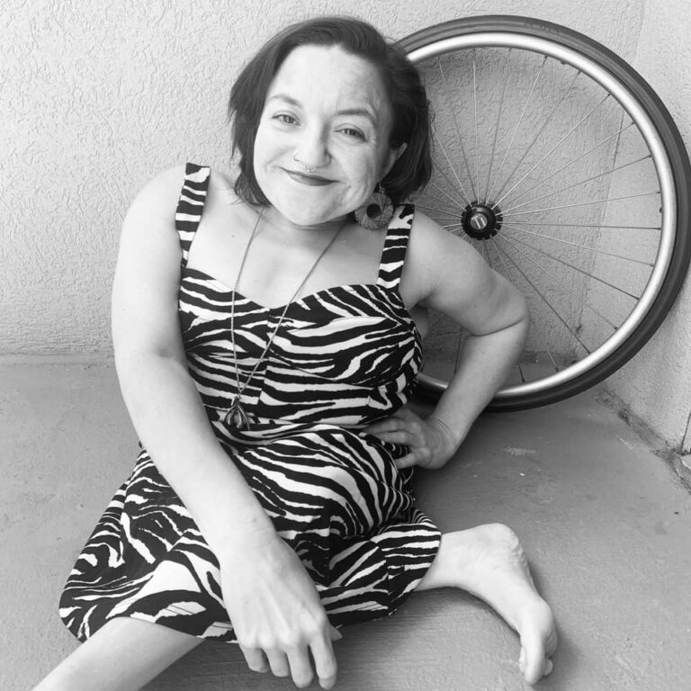 Black and white image of little white woman seated on the floor in a zebra dress with a manual wheelchair wheel behind her.