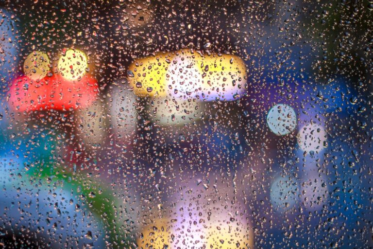 Image of raindrops blurring a glass window, making it difficult to clearly notice what is outside of the window. There appear to be brightly colored signs on the other side of the window, but the view of these signs is quite blurry.
