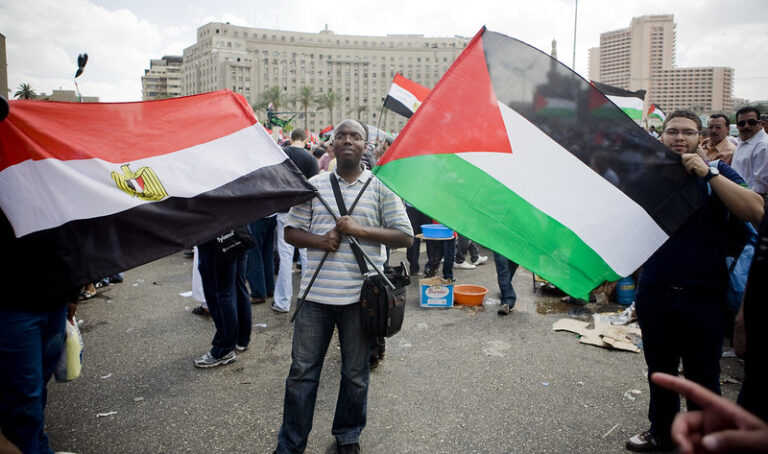 An Egyptian man holds the Egyptian flag in his right hand criss-crossed with the Palestinian flag which he holds in his left hand creating an x on his shirt. He is standing in the middle of Tahrir Square in Cairo with many people in the background but you can't see their faces.