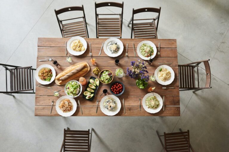 A long wooden table with seven empty chairs, place settings, food, drinks, and decorative flowers.