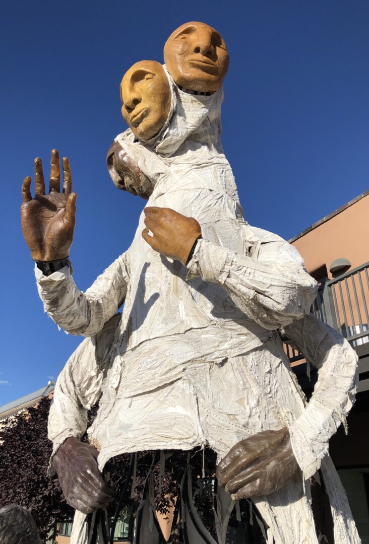 Photo of a 21-foot sculpture outdoor in Santa Fe, New Mexico titled "The Solacii" by Tigre Mashaal-Lively (they/them/their). It has three multi-colored heads and four hands draped in white cloth. It is composed of steel, resin, lights, sound, and donated and reclaimed textiles.