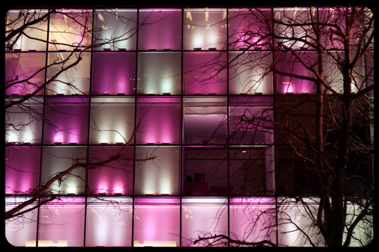 Photo of a seemingly empty office building at night with the windows illuminated by purple, pink, and white lights.