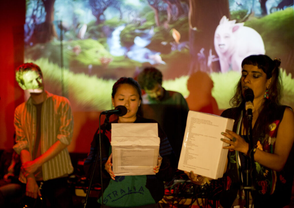 Tara Fatehi and Jemima Yong each holding a white box and speaking into microphones. Pouya Ehsaei playing electronic music. On the back wall projected image of a surreal forest, colourful leaves, a pink pig-like animal.