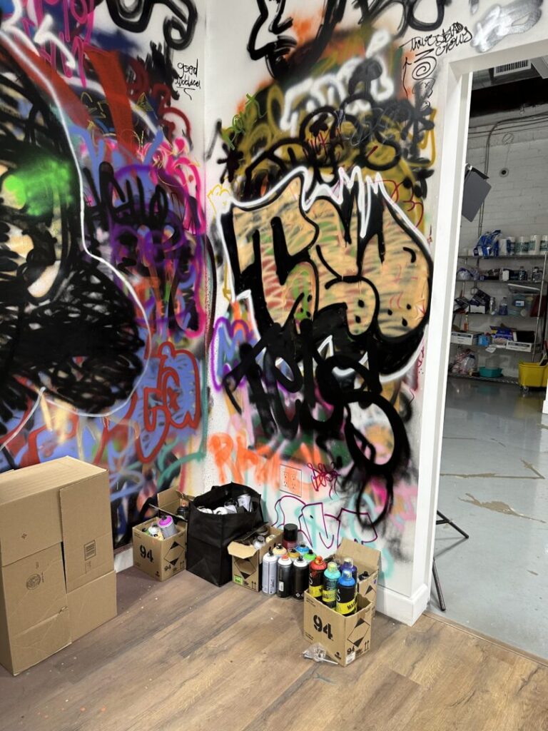 A corner of a white walled room covered in colorful graffiti, including in large, stylized letters the name “TED,” behind cardboard boxes of spray paint cans on a wooden floor. 