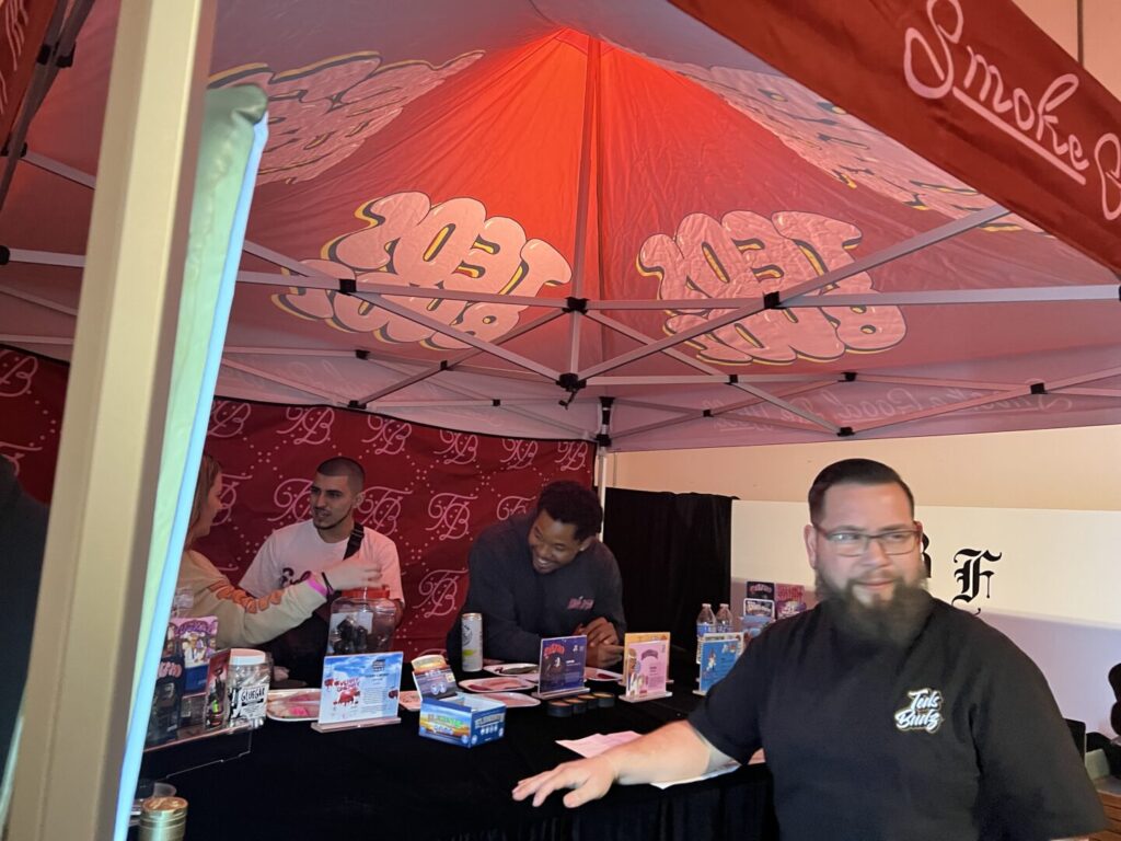 Wearing a black t-shirt with a white Teds Budz logo, Anthony Garcia stands in front of a Teds Budz booth at a dispensary event, cannabis products visible behind him.