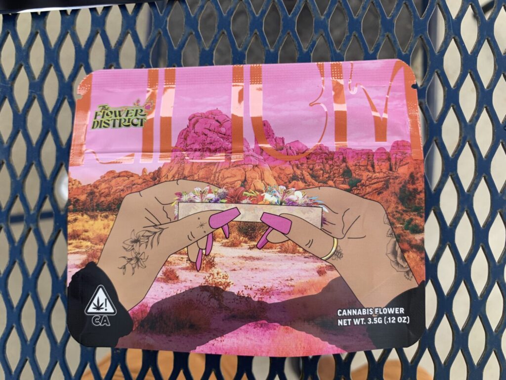 The “Dahlia” strain package depicts two brown, tattooed hands with long pink nails rolling a joint of brightly colored flowers, a pink dessert landscape in the distance.