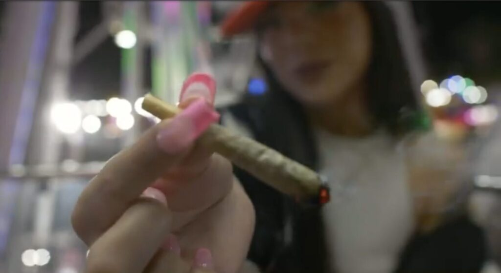 With carnival lights in the background, a young brown woman with long brown hair offers to the viewer the joint she holds between fingers with long pink acrylic nails. 