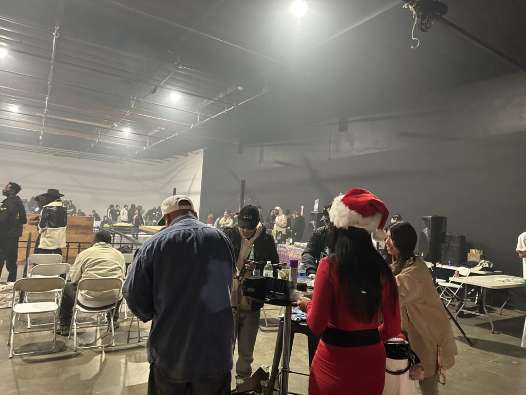 In a large, smokey room, four cannabis enthusiasts, including one dressed as a Christmas elf, surround a table as they smoke, other event attendees visible in the background.