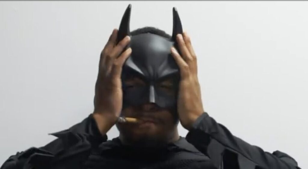 Lewis adjusts his Batman cowl, a lit joint between his lips.