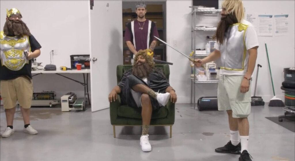 In a white warehouse room with cleaning supplies in the background, Lewis sits in a chair wearing a long brown beard and wig, and a Burger King crown, surrounded by three workers also in fake beards, wigs, and crowns.