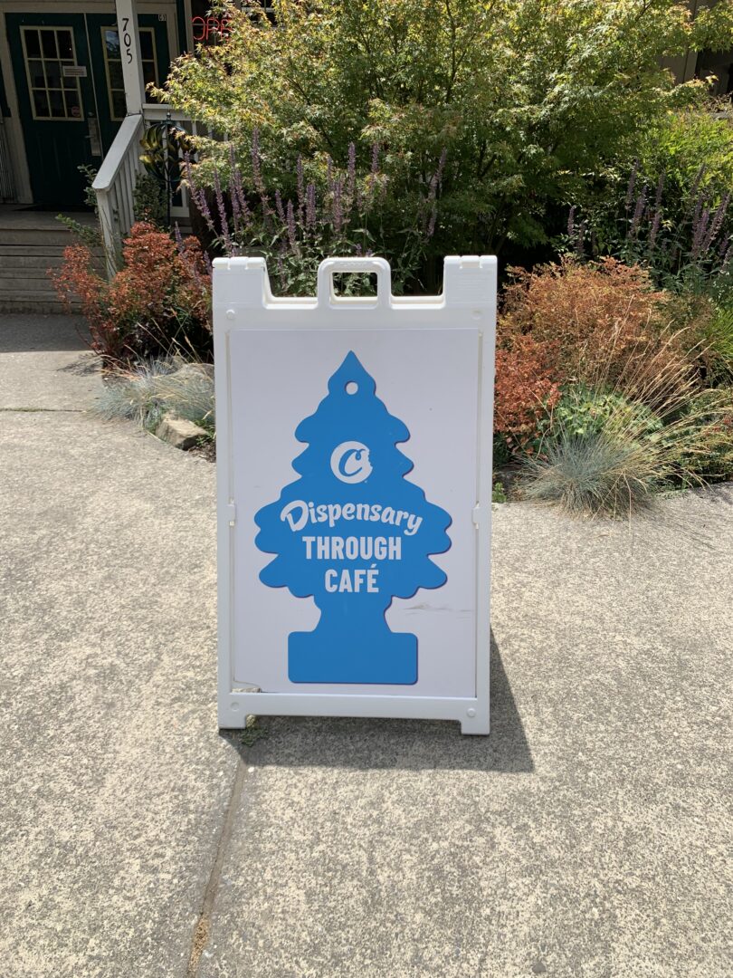 A baby blue sign for the One Log Cookies Dispensary in the shape of a pine tree car air
freshener and reading “Dispensary Through Café.”