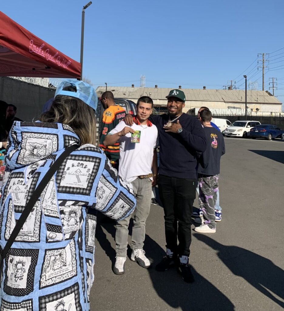 In the foreground left, a figure dressed in blue aims a camera at Theo Lewis and a fan posing in a parking lot, an industrial warehouse in the background.