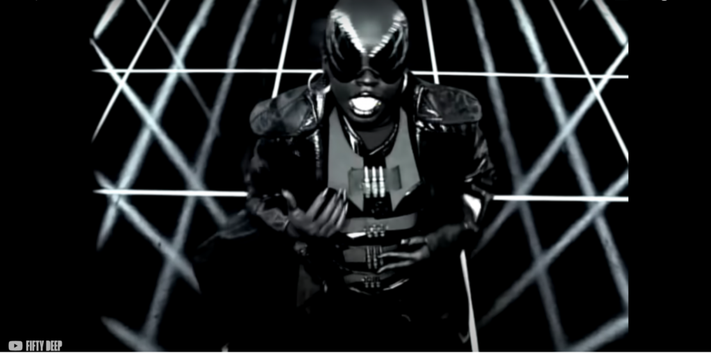 Missy Elliott, a Black woman wearing a shiny black suit and black sunglasses with a mostly black backdrop with white lines coursing through.