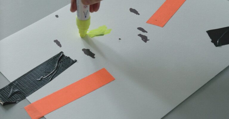 A hand drawing a yellow line on white paper with a large marker. Black and orange lines/shapes in background.