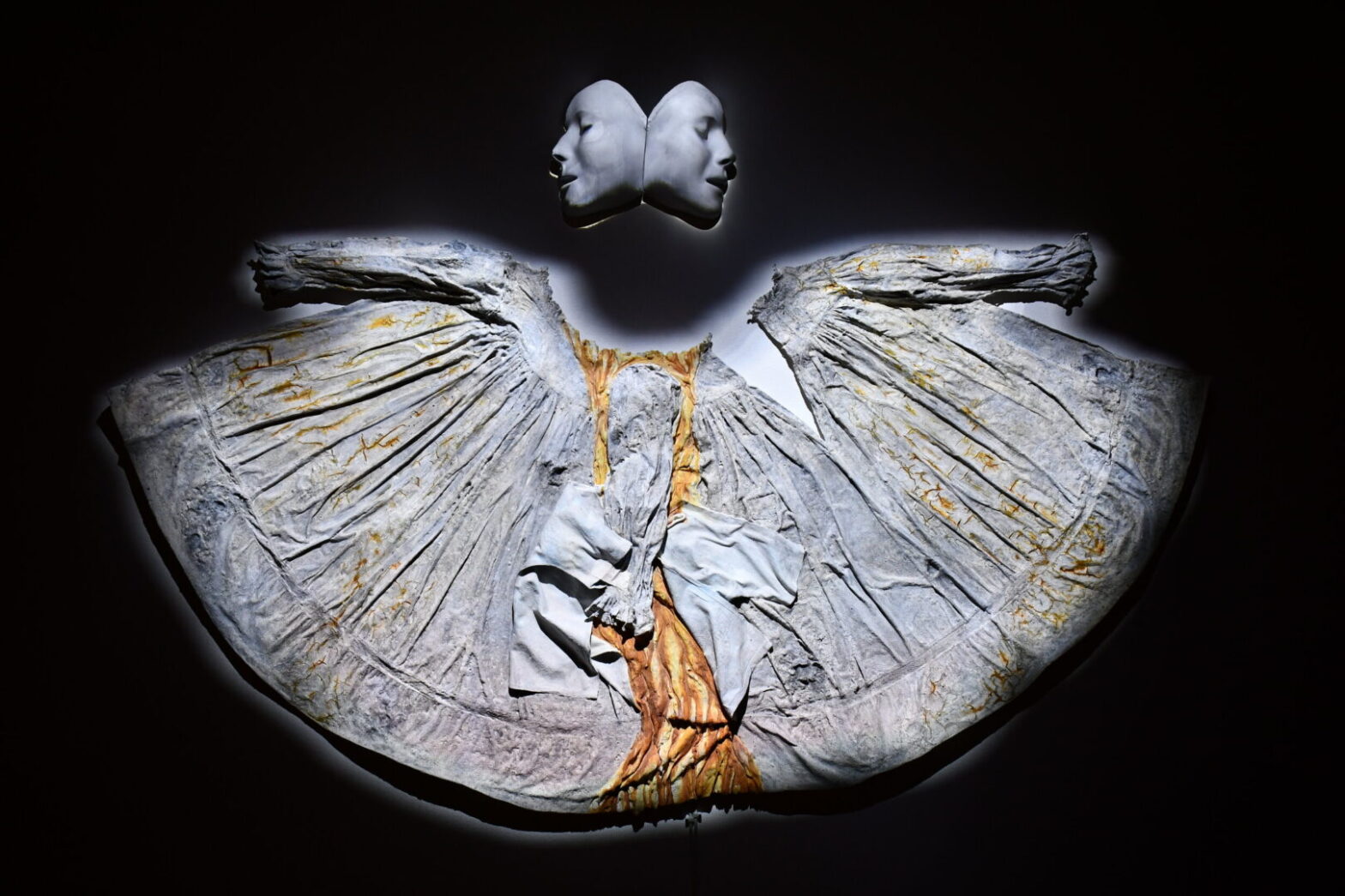 A medicalized undead two-faced figure, facing forwards and looking back, navigates past and future endings from the position of present doom. Made from a baby dress, infant hospital gown, plaster casts of CPR dummy face, plaster, clay, glue, ash, iodine, and plaster bandages.