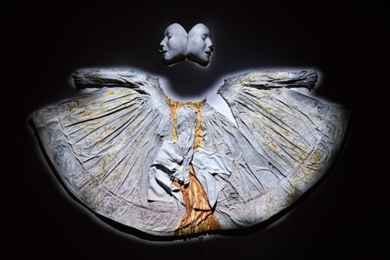 A medicalized undead two-faced figure, facing forwards and looking back, navigates past and future endings from the position of present doom. Made from a baby dress, infant hospital gown, plaster casts of CPR dummy face, plaster, clay, glue, ash, iodine, and plaster bandages.