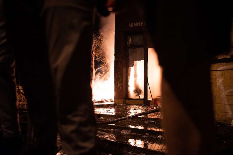 A nighttime photograph taken through the silhouettes of people in the foreground, looking toward a building engulfed in flames. Bright orange fire glows from within the glass doorway and windows of a dark structure, with the word "MINNEA" visible above the entrance. Smoke and embers rise from a separate fire burning next to a tree to the left of the building. The ground in the foreground is wet and littered with debris, reflecting the intense light from the fires.