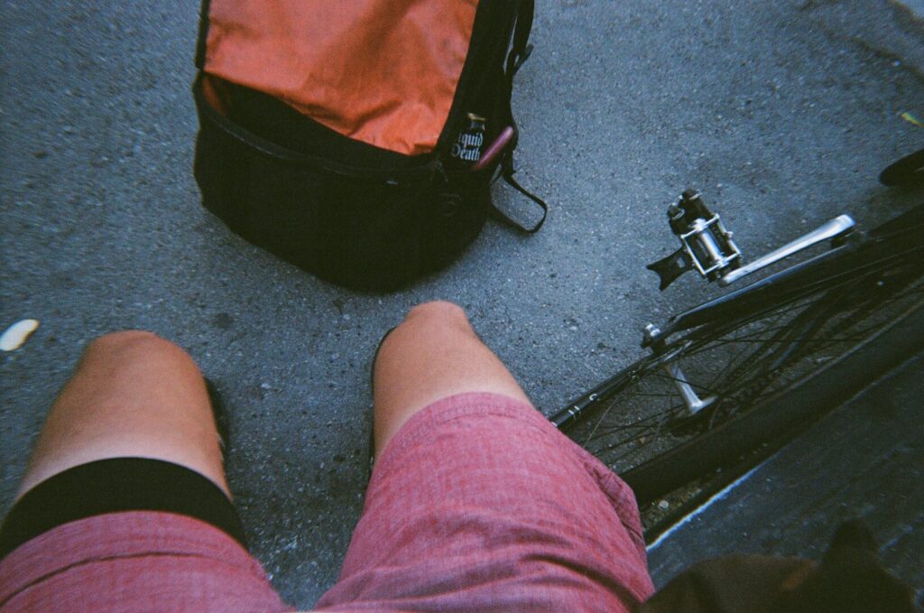 Picture taken from the point of view of a rider. Shorts reveal tanned legs. On the ground next to him we see a bicycle and a large backpack.